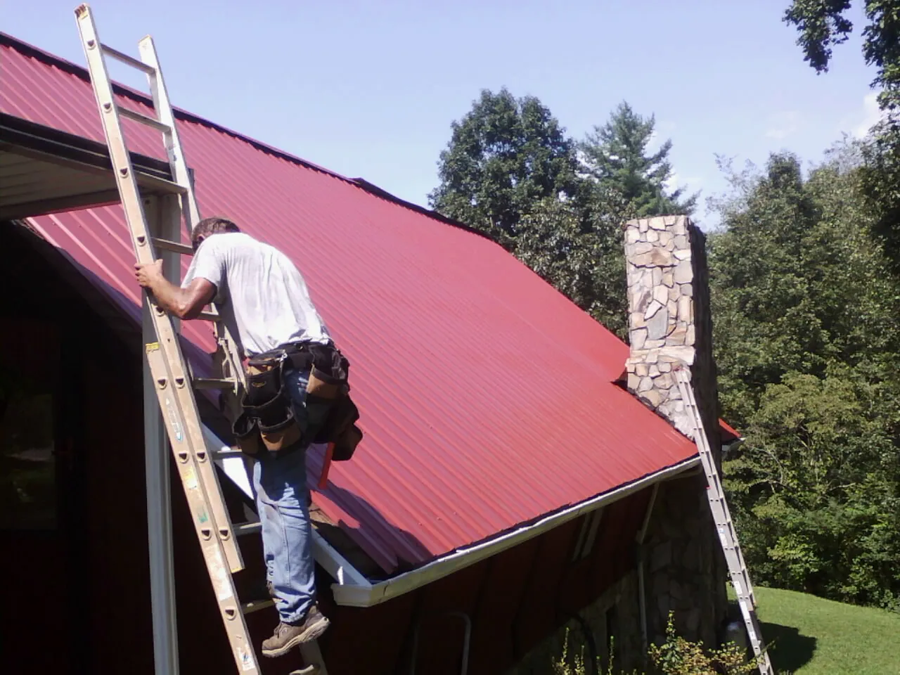 Roofer on a ladder beside a red metal roof and stone chimney — Soffit & Fascia Repair by Roofing Brothers in Crockett
